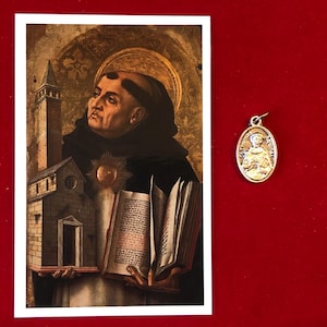 May include: A vintage-style religious image of a man in a brown robe holding a book and a small model of a church. The image is framed in a white border. A silver oval pendant with a religious image is shown to the right of the image.