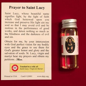 Op de afbeelding: Een witte kaart met een gebed tot Sint Lucia, een klein flesje olie met een wit etiket waarop een afbeelding van Sint Lucia staat, en de tekst "Geraakt aan een relikwie van: Sint Lucia, Maagd en Martelaar".