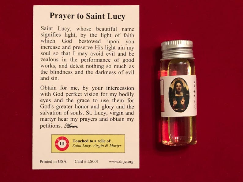 Op de afbeelding: Een witte kaart met een gebed tot Sint Lucia, een klein flesje olie met een wit etiket waarop een afbeelding van Sint Lucia staat, en de tekst "Geraakt aan een relikwie van: Sint Lucia, Maagd en Martelaar".