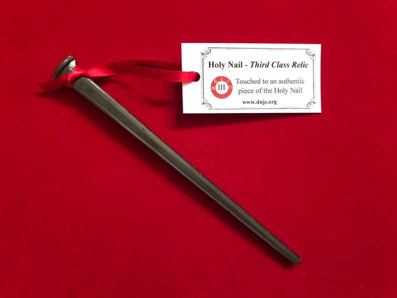 May include: A black metal nail with a red ribbon tied around it. A white card with the text "Holy Nail - Third Class Relic" and "Touched to an authentic piece of the Holy Nail" and "www.dnjc.org" is attached to the ribbon.