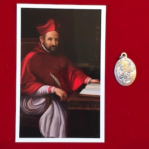 May include: A vintage-style religious card featuring a painting of a man in a red robe and a white hat. The man is holding a quill pen and a book. The card is next to a silver oval-shaped medal with the words "St. Robert Bellarmine" engraved on it.