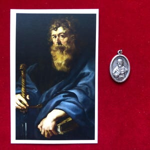 Könnte beinhalten: Eine religiöse Karte mit einem Gemälde eines bärtigen Mannes, der ein Schwert und ein Buch hält. Neben der Karte befindet sich ein ovaler Anhänger aus Silber, der einen Mann mit einem Schwert darstellt.