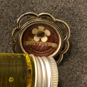Puede incluir: Un colgante reliquia de tono plateado con una ampolla de vidrio que contiene una peque&ntilde;a flor blanca y un trozo de tela con el texto "S. Teresiae a I. I."