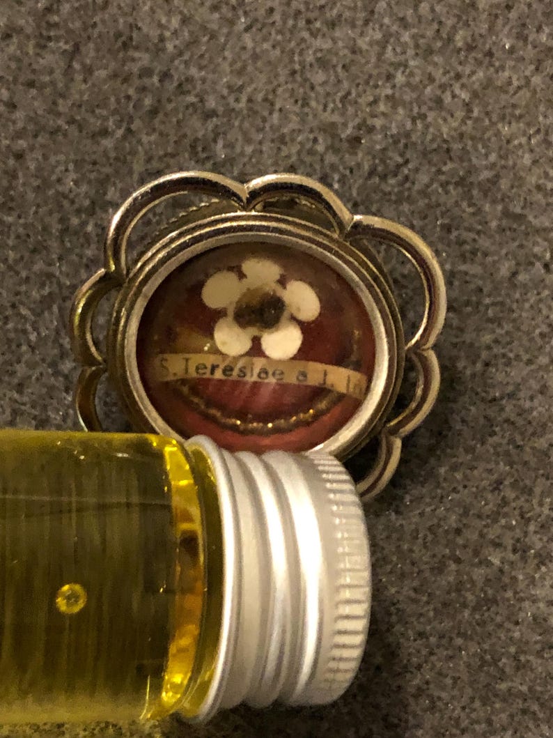 Puede incluir: Un colgante reliquia de tono plateado con una ampolla de vidrio que contiene una peque&ntilde;a flor blanca y un trozo de tela con el texto "S. Teresiae a I. I."