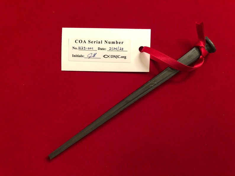 May include: A black metal nail with a COA (Certificate of Authenticity) tag attached. The tag has the text "COA Serial Number" and the number "N23-001". The date is "2/24/23" and the initials "JB" are written on the tag. The tag also has the website "DNJC.org".