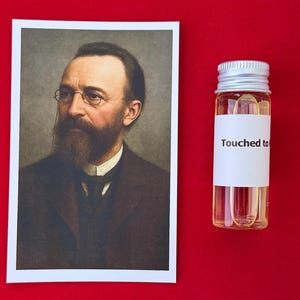 以下が含まれることがあります： 長いひげと眼鏡をかけた男性の肖像画と、銀色のキャップと「Touched to」と書かれた白いラベルが付いた小さなガラスバイアル。背景は赤色です。
