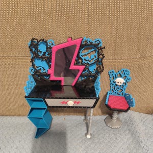 May include: A black and blue toy vanity set with a pink lightning bolt-shaped mirror. The set includes a matching chair with a skull design and a small blue shelf. The vanity has a silver leg.