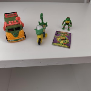 Pode incluir: Uma coleção de brinquedos Tartarugas Ninja, incluindo uma carrinha amarela e verde, uma mota amarela e verde, uma figura de ação verde e uma pequena banda desenhada com Rafael.