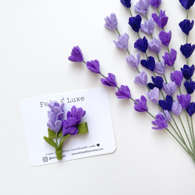 May include: A felt lavender flower hair clip with a green stem. The clip is on a white card with the text "Felt & Luxe" and the website address "www.feltandluxe.etsy.com".