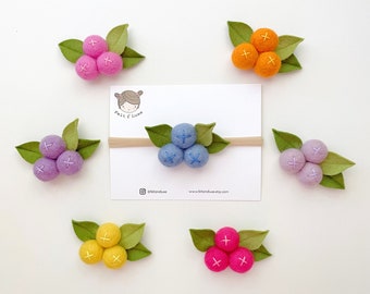 May include: A collection of six felt berry hair accessories in pink, orange, purple, light blue, yellow, and magenta, each featuring three spheres with embroidered cross details and green felt leaves. One blue berry accessory is attached to a beige headband.