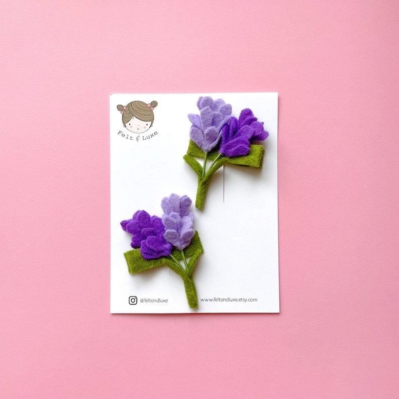 May include: Two felt flower hair clips in shades of purple and green. The flowers are arranged in a bouquet shape and are attached to a green felt stem. The hair clips are labeled "Felt & Luxe" and have the website address "www.feltandluxe.etsy.com".