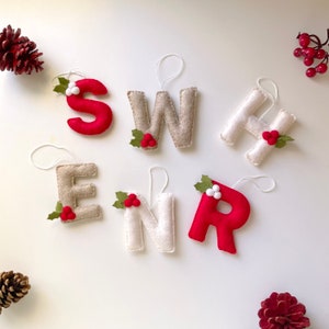May include: Six felt letter ornaments in red, white, and beige, each featuring a white hanging string and a small decorative holly sprig with red or white berries. The letters spell S, W, H, E, N, and R, arranged in two rows with pinecones.