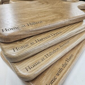 K&ouml;nnte beinhalten: Ein Stapel Holzschneidebretter mit Griffen. Jedes Brett hat eingravierten Text, darunter "Home at Hillcot" und "Home with the Hares". Die Bretter haben eine nat&uuml;rliche Holzmaserung und eine helle Holzfarbe.