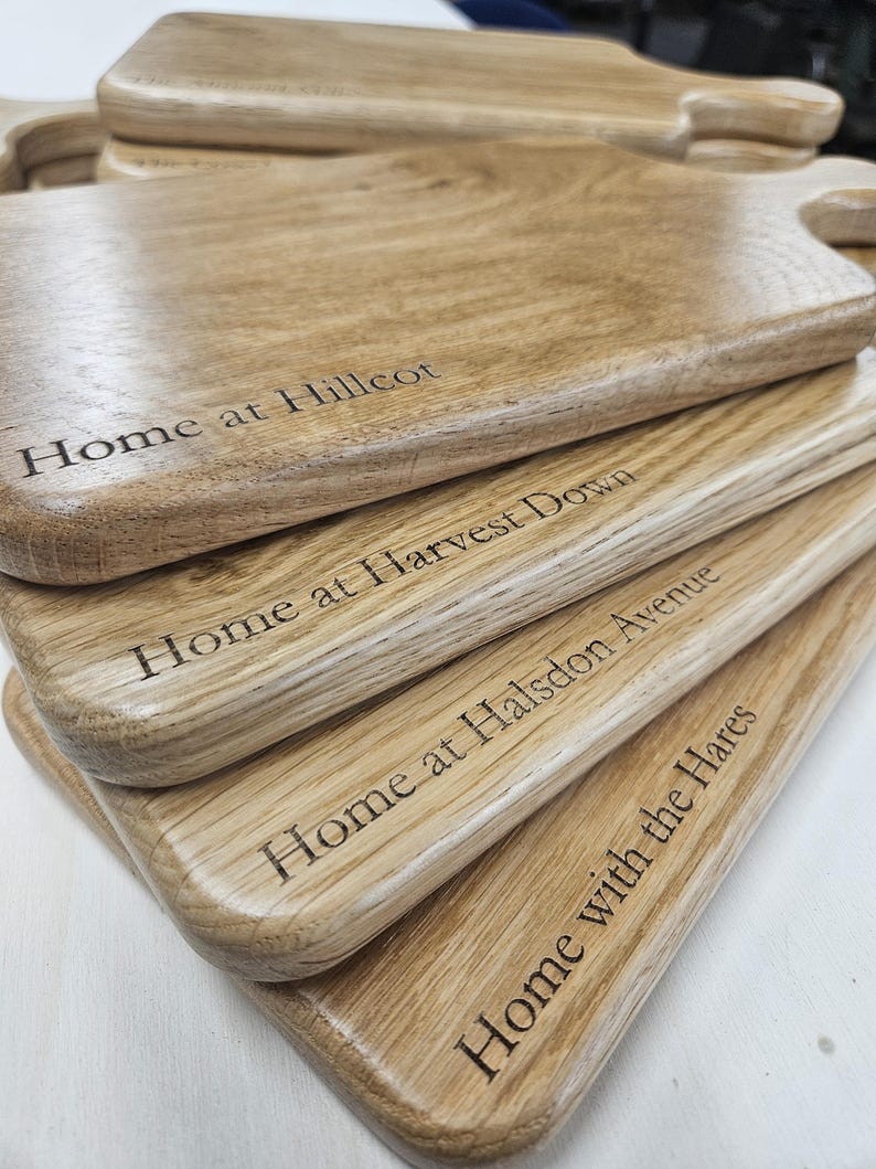 K&ouml;nnte beinhalten: Ein Stapel Holzschneidebretter mit Griffen. Jedes Brett hat eingravierten Text, darunter "Home at Hillcot" und "Home with the Hares". Die Bretter haben eine nat&uuml;rliche Holzmaserung und eine helle Holzfarbe.
