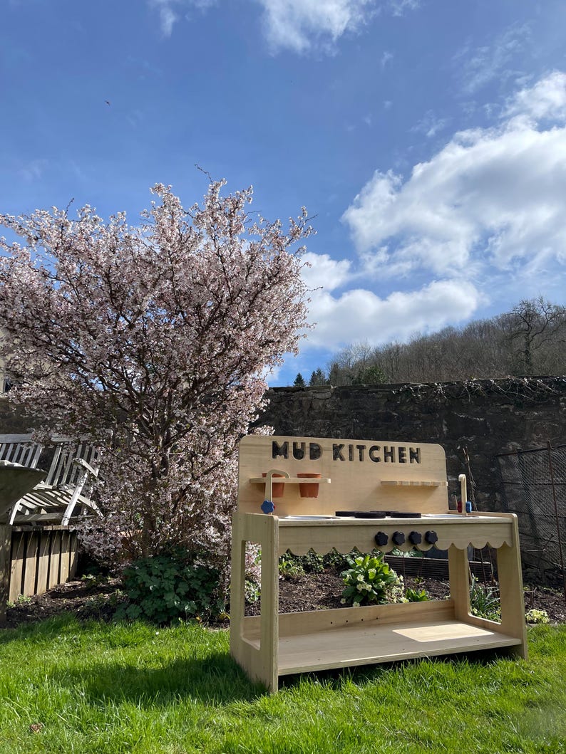 May include: A wooden outdoor mud kitchen playset with a sign that reads "MUD KITCHEN". The playset has a sink, a stovetop, and shelves. It is set up in a grassy backyard with a flowering tree in the background.
