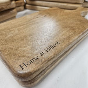 K&ouml;nnte beinhalten: Gestapelte Holzschneidebretter mit Griffen. Die Bretter sind hellbraun mit sichtbarer Holzmaserung und abgerundeten Kanten. Der Schriftzug "Home at Hillcot" ist auf der Vorderseite eingraviert.