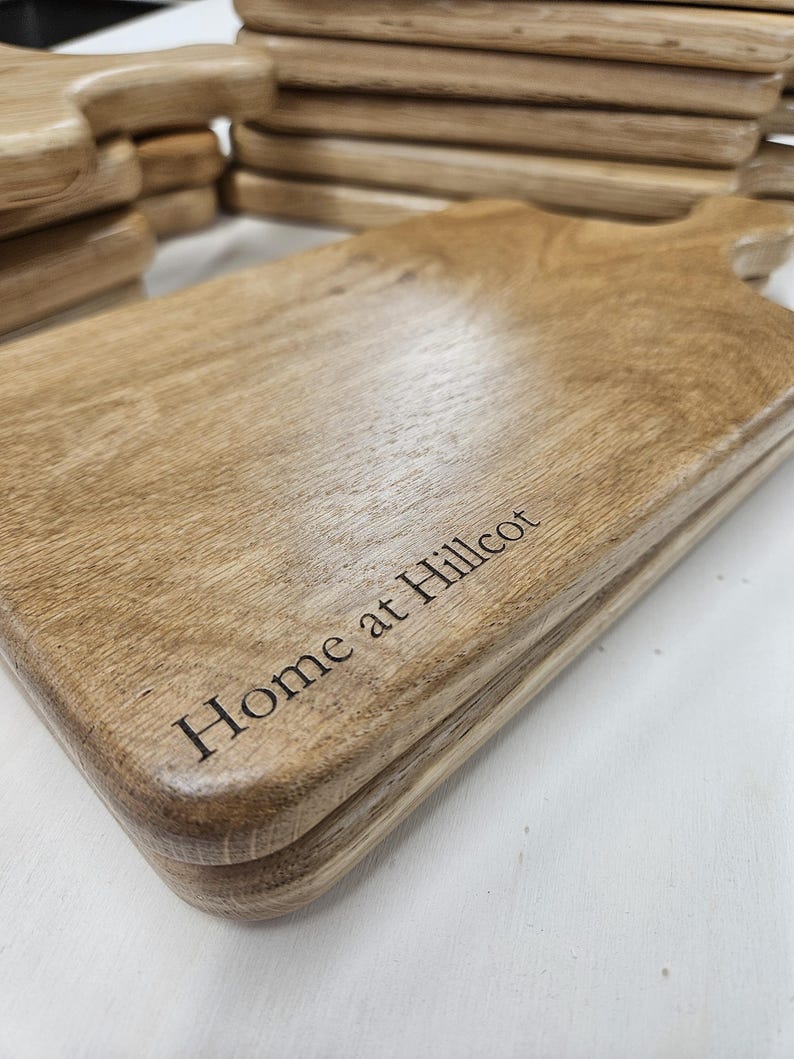 K&ouml;nnte beinhalten: Gestapelte Holzschneidebretter mit Griffen. Die Bretter sind hellbraun mit sichtbarer Holzmaserung und abgerundeten Kanten. Der Schriftzug "Home at Hillcot" ist auf der Vorderseite eingraviert.
