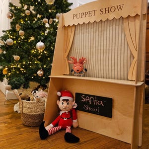 May include: A wooden puppet show stage with the words "PUPPET SHOW" engraved on top. A plush elf in a Santa hat and a reindeer puppet are inside. A small chalkboard reads "SANTA Show". A Christmas tree is in the background.
