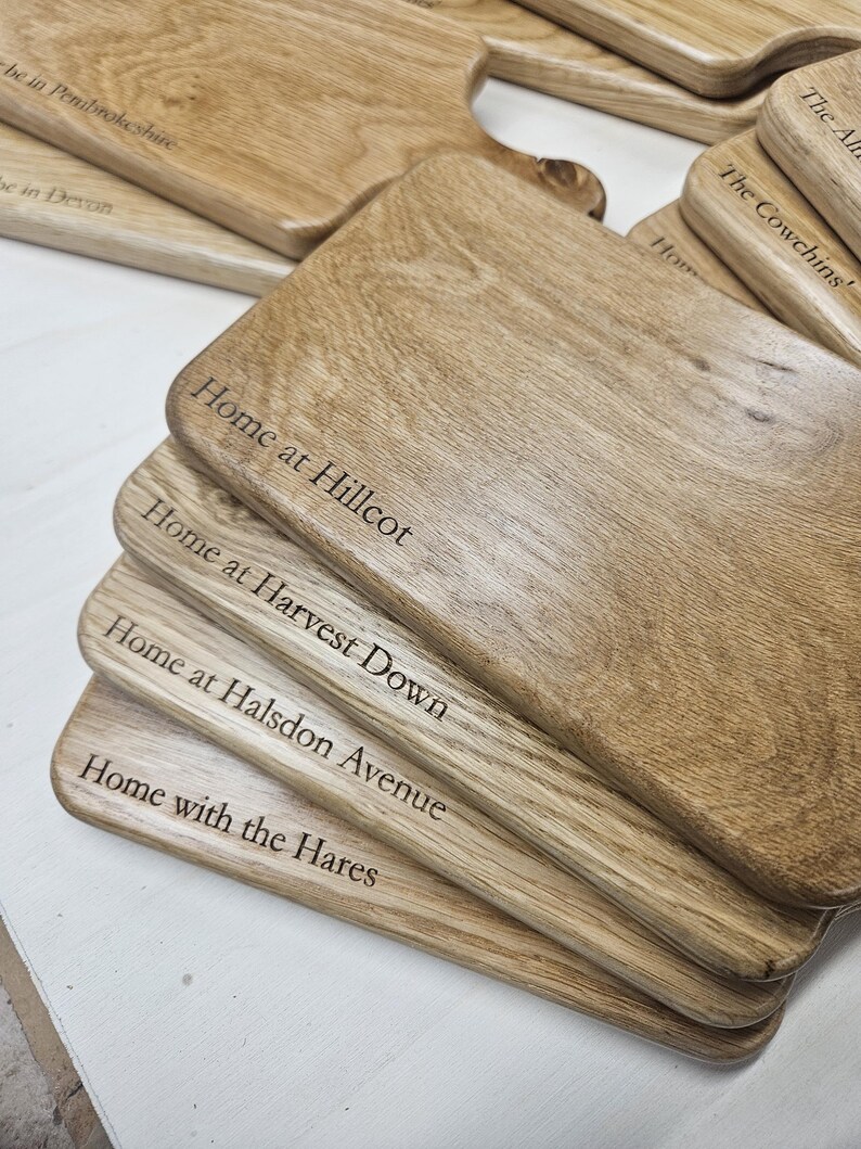 K&ouml;nnte beinhalten: Eine Reihe von Holzschneidebrettern mit Gravuren. Die Bretter sind hellbraun und zeigen eine deutliche Holzmaserung. Texte umfassen "Home at Hillcot", "Home at Harvest Down", "Home at Halsdon Avenue" und "Home with the Hares".