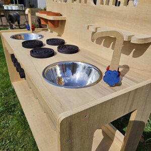 May include: A wooden mud kitchen playset with a stainless steel sink, a wooden faucet, and a stovetop with four black burner covers. The kitchen has a shelf for storage and the words "MUD KITCHEN" are cut out of the back panel.