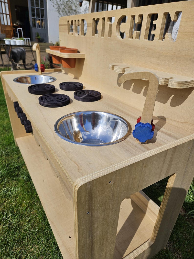 May include: A wooden mud kitchen playset with a stainless steel sink, a wooden faucet, and a stovetop with four black burner covers. The kitchen has a shelf for storage and the words "MUD KITCHEN" are cut out of the back panel.