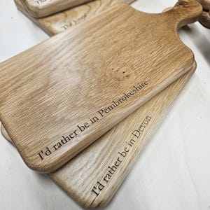 K&ouml;nnte beinhalten: Mehrere Holzschneidebretter mit Griffen sind gestapelt. Die Bretter haben eine hellbraune Farbe und eine nat&uuml;rliche Holzmaserung. Auf einem Brett steht "I'd rather be in Pembrokeshire", auf einem anderen "I'd rather be in Devon".