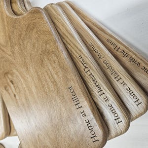 K&ouml;nnte beinhalten: Eine Sammlung von Holzschneidebrettern mit Griffen, mit graviertem Text. Die Bretter sind hellbraun mit sichtbarer Holzmaserung. Der Text enth&auml;lt S&auml;tze wie "Home at Hillcot" und "Home with the Hares."