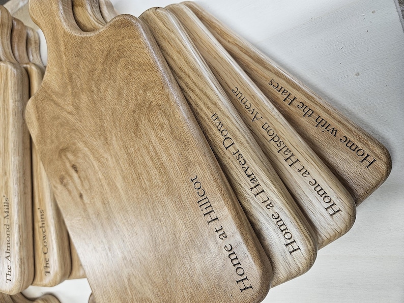 K&ouml;nnte beinhalten: Eine Sammlung von Holzschneidebrettern mit Griffen, mit graviertem Text. Die Bretter sind hellbraun mit sichtbarer Holzmaserung. Der Text enth&auml;lt S&auml;tze wie "Home at Hillcot" und "Home with the Hares."