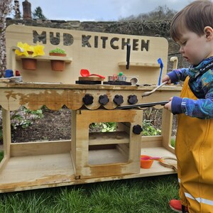 May include: A wooden mud kitchen playset with a sign that reads "MUD KITCHEN". The playset has a stovetop with four knobs, a sink, and shelves. A young child is playing with the kitchen.