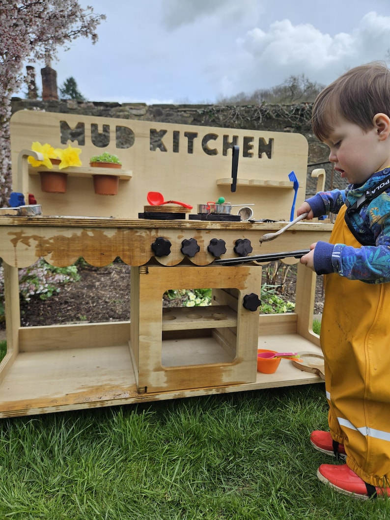 May include: A wooden mud kitchen playset with a sign that reads "MUD KITCHEN". The playset has a stovetop with four knobs, a sink, and shelves. A young child is playing with the kitchen.