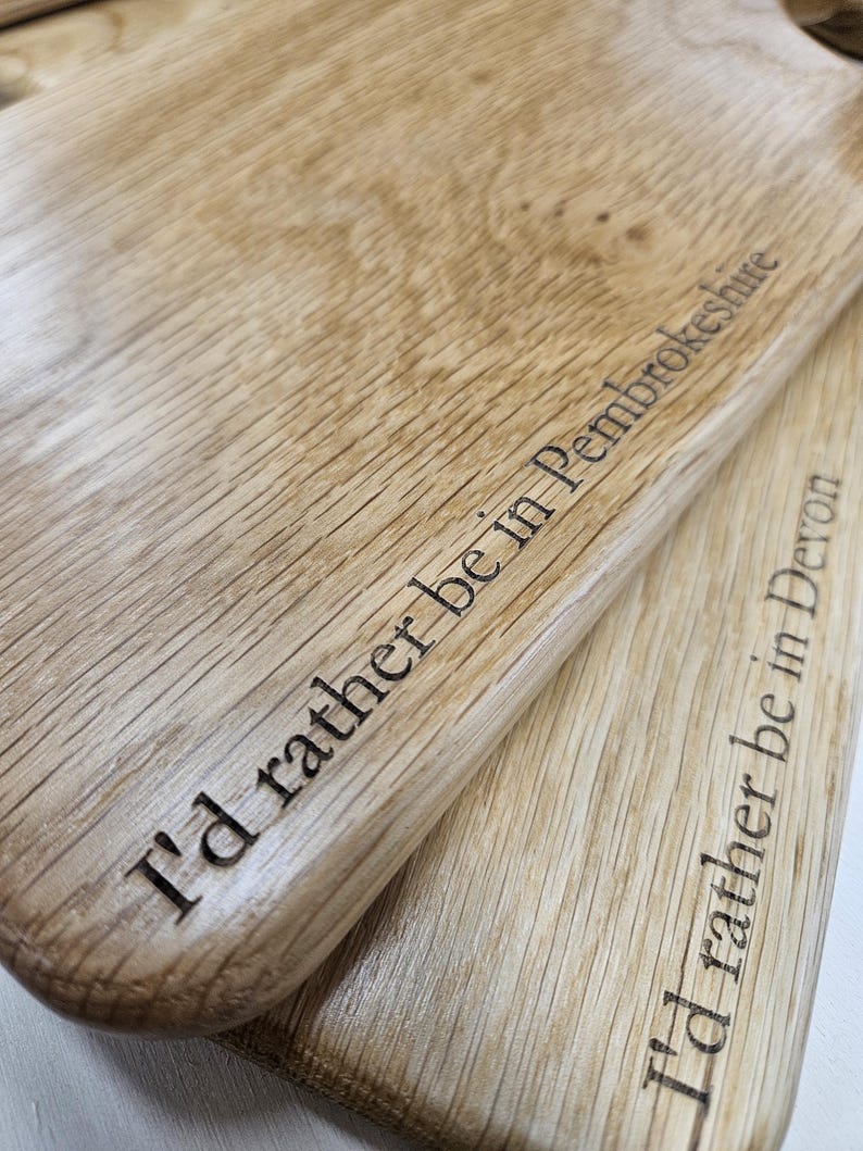 K&ouml;nnte beinhalten: Zwei helle Holzschneidebretter mit den eingravierten Texten "I'd rather be in Pembrokeshire" und "I'd rather be in Devon". Die Bretter haben eine nat&uuml;rliche Holzmaserung.