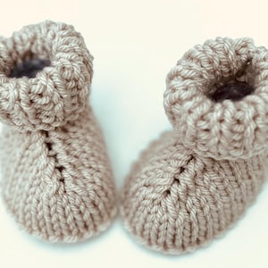 Könnte beinhalten: Ein Paar beige gestrickte Babyschuhe mit einfachem Design. Die Schuhe sind aus weichem Garn gefertigt und haben eine bequeme Passform.
