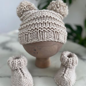 May include: A beige knitted hat with two pom-poms on top and a pair of matching beige knitted booties. The hat features a textured ribbed pattern around the base and a stockinette stitch body. The booties have a folded cuff and a central seam.