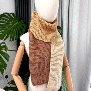May include: A chunky knit scarf featuring a colour-blocked design with three distinct sections in shades of tan, medium brown, and cream. The scarf is wrapped around the neck of a mannequin, displaying a consistent garter stitch texture throughout.