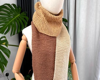 Può includere: Una sciarpa in maglia grossa caratterizzata da un design a blocchi di colore con tre sezioni distinte nelle tonalità marrone chiaro, marrone medio e crema. La sciarpa è avvolta attorno al collo di un manichino e presenta una trama a legaccio.