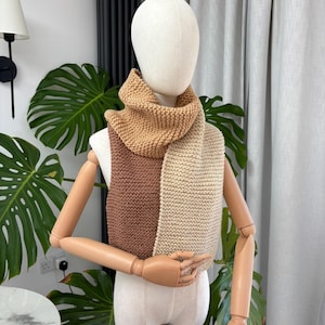 Könnte beinhalten: Ein gestrickter Schal mit einem Color-Block-Design in drei verschiedenen Braun- und Beigetönen. Der Schal ist im kraus rechts Muster gestrickt und zeigt einen dunkelbraunen Bereich, einen helleren beigen Bereich und einen warmen kamelfarbenen Kragen.