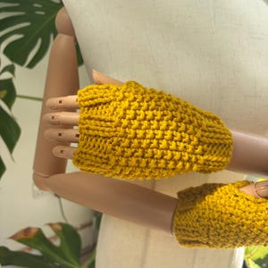 May include: A pair of mustard yellow fingerless gloves featuring a textured crochet stitch pattern. The gloves have a ribbed cuff design at the wrist and a separate opening for the thumb, crafted from a thick yarn with a visible knit-like texture.