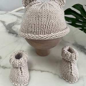 May include: A light beige knitted hat with two small rounded ears on top and a matching pair of knitted booties. The hat features a ribbed texture and a rolled brim, while the booties have a thick, cuffed opening and a textured knit pattern.