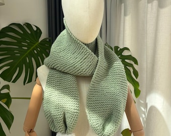 May include: A sage green knitted scarf featuring a garter stitch texture and two white pom-poms attached to the ends. The scarf is draped around the neck of a neutral-toned mannequin, showcasing the soft yarn construction and the rounded pom-pom details.