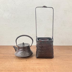 以下が含まれることがあります： 湾曲したハンドルと質感のある表面を持つダークブラウンの鋳鉄製ティーポットと、高く細い金属製ハンドルを備えた背の高い正方形の編み込み竹製バスケットが、平らな表面に並べて置かれています。