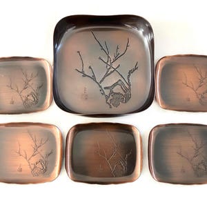 以下が含まれることがあります： 銅色の金属製トレイ6個セット。大きな正方形のトレイ1つと、小さな長方形のトレイ5つが含まれています。各トレイには、葉と岩のある枝のエッチングデザインが施されています。トレイには濃い茶色の縁があります。
