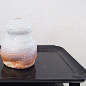 以下が含まれることがあります： 黒いトレイの上に置かれた、白地に茶色の釉薬がかかった陶器の花瓶。花瓶は丸みを帯びた球根状で、開口部はわずかに広がっています。