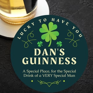 May include: A dark green round coaster with the text "LUCKY TO HAVE YOU" in a white cursive font. The coaster also features a green four-leaf clover, two smaller green clovers, and the text "DAN'S GUINNESS" in a white font. The text "A Special Place, for the Special Drink of a VERY Special Man" is also included in a white font.