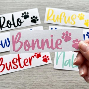 May include: White rectangular stickers with pet names in various colours, including black, yellow, blue, red, and pink. Each sticker features a paw print design. The name "Bonnie" is in pink, and "Rolo" is in black.