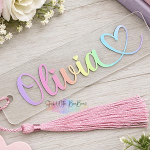 May include: A clear rectangular acrylic bookmark featuring the name Olivia in a gradient rainbow script font with a small heart above the i. A pink cord is attached to the top left corner, leading to a long, matching pink fabric tassel.