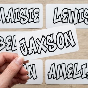 May include: White rectangular stickers with black outlined bubble letters spelling out names: Maisie, Lewis, Bella, Jaxson, and Amelia. One sticker is held by a hand. The stickers are on a light wood surface.