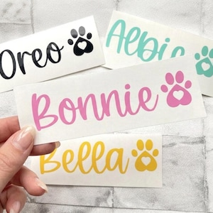 May include: A hand holds several white rectangular stickers with pet names and paw print designs. The names "Oreo" in black, "Bonnie" in pink, "Bella" in yellow, and "Albie" in teal are displayed. Each paw print includes a heart.