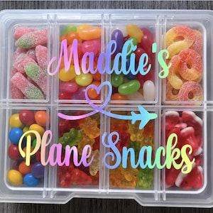 May include: A clear plastic rectangular storage container with eight compartments filled with assorted colourful sweets. The lid features a gradient rainbow-coloured decal with the text Maddie's Plane Snacks and a heart-shaped aeroplane icon.