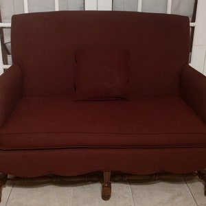 May include: A burgundy loveseat with a matching pillow. The upholstered sofa has a high back and armrests. The wooden legs are a darker brown. The scalloped skirt adds a decorative touch to the furniture.