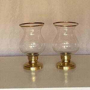 May include: Two clear glass candle holders with gold rims and bases. The candle holders have a bulbous shape and sit on a gold pedestal base. The background is a light beige colour.